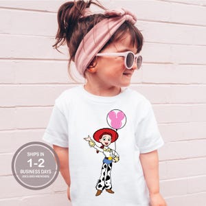 May include: White t-shirt with a cartoon cowgirl character holding a pink balloon. The cowgirl wears a red hat, white shirt, and black and white pants. The shirt has the text "SHIPS IN 1-2 BUSINESS DAYS (EXCLUDES WEEKENDS)" in a gray circle.