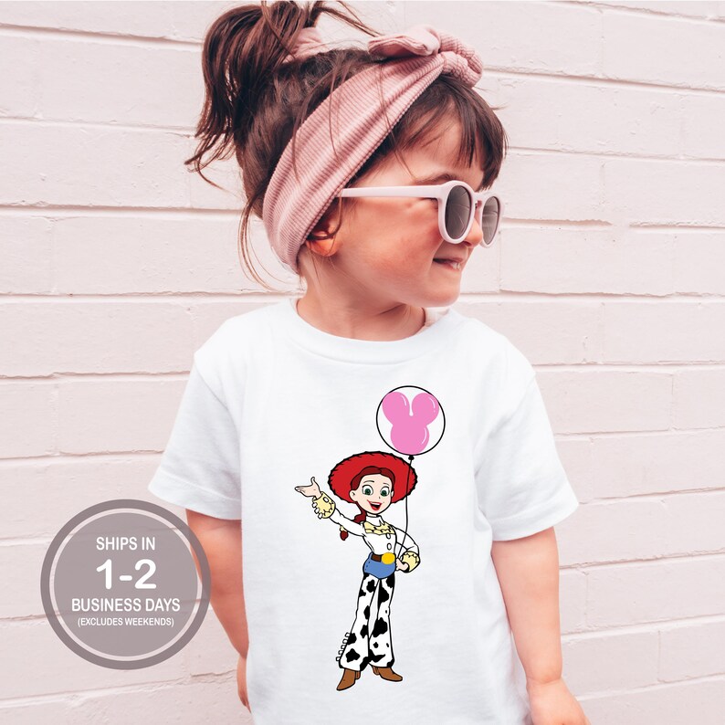 May include: White t-shirt with a cartoon cowgirl character holding a pink balloon. The cowgirl wears a red hat, white shirt, and black and white pants. The shirt has the text "SHIPS IN 1-2 BUSINESS DAYS (EXCLUDES WEEKENDS)" in a gray circle.