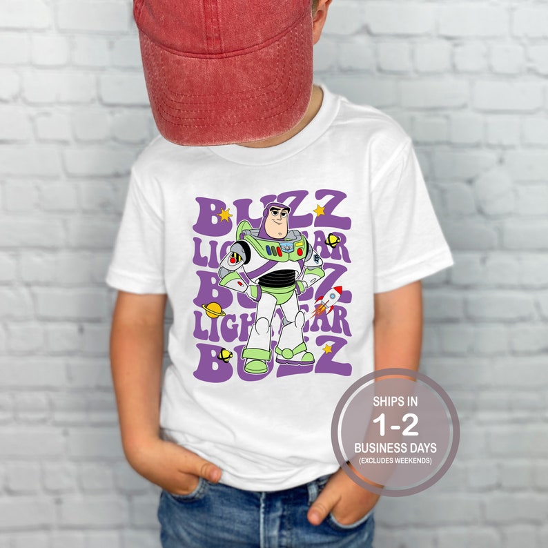 K&ouml;nnte beinhalten: Wei&szlig;es T-Shirt mit einem lila und gr&uuml;nen Aufdruck von Buzz Lightyear aus Toy Story. Der Aufdruck enth&auml;lt den Text "Buzz Lightyear", der in einem Retro-Stil wiederholt wird.