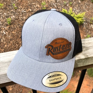 May include: Gray and black baseball cap with a brown leather patch that says "Medicated Rust-eze Bumper Ointment Rear End Formula". The cap also has a gold sticker that says "The Authentic Yupoong Snapback Yupoong Since 1974".