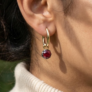 May include: Gold hoop earrings with a dangling, round, deep red gemstone. The earrings are worn on the ear of a person wearing a cream-colored turtleneck sweater. The background is blurred, with green foliage visible.