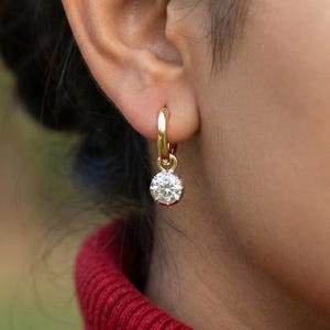 May include: A gold hoop earring with a round, clear gemstone. The earring is hanging from a person's ear. The person is wearing a red sweater. The background is blurred.