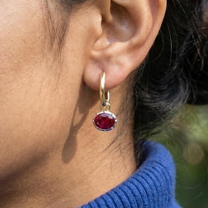 May include: Gold hoop earrings with oval, deep red gemstones. The gemstones are set in a silver-colored bezel. The earrings are shown on a person's ear, with a blue sweater in the background.