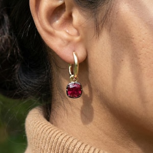 May include: Gold hoop earrings with a square, deep red gemstone pendant. The earrings are worn on a person's ear, with the focus on the jewelry. The background is a blurred green and brown.