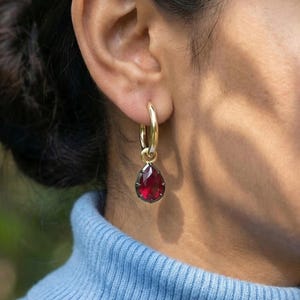 May include: A gold hoop earring with a teardrop-shaped, deep red gemstone pendant. The earring is worn on a person's ear, with the focus on the jewelry. The gemstone is faceted, catching the light and creating a sparkling effect. The earring is a classic and elegant accessory.