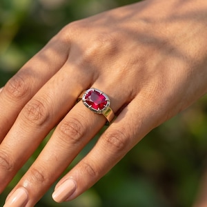 Puede incluir: Un anillo con una gran piedra preciosa de color rojo intenso, tallada en forma rectangular y engastada en una banda de metal plateado pulido. La piedra está asegurada por cuatro garras prominentes en las esquinas, creando un diseño clásico.