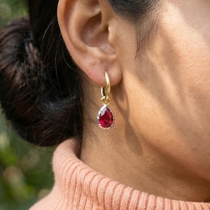 May include: Gold hoop earrings with a teardrop-shaped, deep red gemstone pendant. The earrings are worn on the ear of a person wearing a peach-colored turtleneck sweater. The background is blurred, with green foliage.