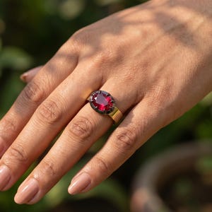 May include: A ring featuring a large, oval-cut, deep red gemstone set in a dark, metallic prong setting. The band of the ring is a smooth, polished gold-tone metal that contrasts with the dark setting of the central red stone.