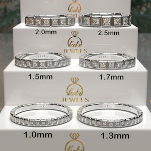 Op de afbeelding: Vijf zilveren ringen met vierkante diamanten in een rij. De ringen worden gepresenteerd op een witte, meerlagige standaard. De ringen zijn gelabeld met hun breedte in millimeters: 1,0 mm, 1,3 mm, 1,5 mm, 1,7 mm en 2,0 mm.