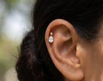 May include: A gold cartilage earring featuring a small round ball stud connected to a dangling clear pear-shaped gemstone. The jewelry is positioned in the upper ear cartilage, showcasing a polished gold metal finish and a faceted white stone.