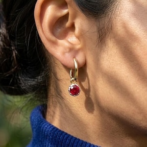 May include: A close-up of a gold hoop earring with a round, red gemstone. The earring is hanging from a person's ear. The person is wearing a blue turtleneck sweater. The background is blurred.