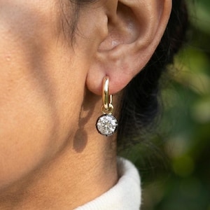 Puede incluir: Primer plano de un pendiente de aro dorado con una gema redonda y transparente. La gema está engastada en un bisel de metal oscuro. El pendiente está sujeto a una oreja.