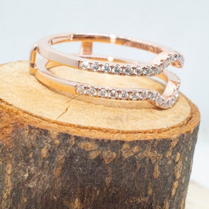 Puede incluir: Un anillo de oro rosa con un dise&ntilde;o de doble banda. La banda superior est&aacute; engastada con peque&ntilde;os diamantes y tiene una forma curva. El anillo se muestra sobre una superficie de madera.