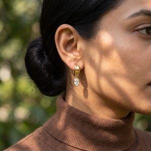May include: A gold hoop earring featuring a polished, double-banded design at the top. A single, clear, oval-shaped gemstone is suspended from the centre of the hoop, secured by a smooth gold bezel setting that catches the light.