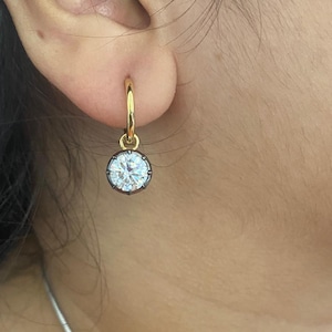 May include: A gold hoop earring with a round, clear gemstone pendant. The gemstone is set in a dark metal bezel. The earring is worn in the ear of a person.