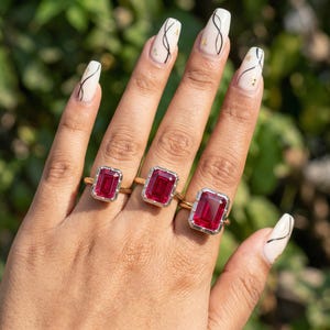 Könnte beinhalten: Drei goldene Ringe mit großen, rechteckigen, rubinroten Edelsteinen. Die Ringe sind in Gold gefasst und haben einen silberfarbenen Rand um die Edelsteine. Die Ringe befinden sich an einer Hand mit langen, gepflegten Nägeln.