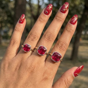 May include: Three gold rings with large, square, ruby-red gemstones are displayed on a hand. The rings have a simple, elegant design. The nails are painted red with gold accents.
