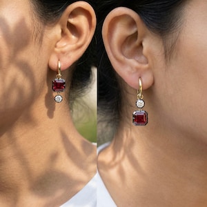 May include: Gold hoop earrings with a square, deep red gemstone and a smaller, round, white gemstone dangling below. The earrings are set against a blurred background, highlighting the jewelry's design and color.