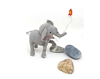 May include: A gray crocheted elephant toy with white tusks and pink cheeks, holding a white string with a red and orange flame at the end. The elephant is standing on a white surface with three gray and brown rocks.