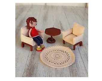 May include: A crocheted doll sitting in a crocheted armchair with a crocheted coffee table and a crocheted rug. The doll is wearing a red shirt and blue pants.
