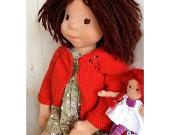 May include: A handmade cloth doll with brown yarn hair, wearing a red knitted cardigan over a green floral dress. A smaller doll with red hair and a white dress is also present. The dolls are sitting on a stack of books.