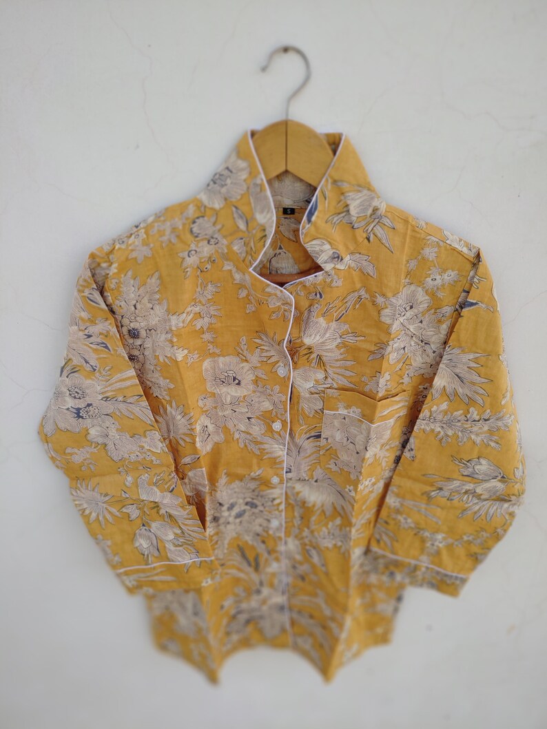 May include: A yellow button-up shirt with a white floral print and white trim. The shirt has a mandarin collar and long sleeves.