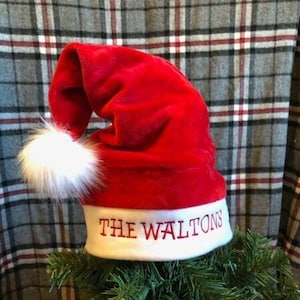 May include: Red velvet Santa hat with a white faux fur trim and the words "The Waltons" embroidered on the brim. The hat is sitting on top of a green Christmas tree.