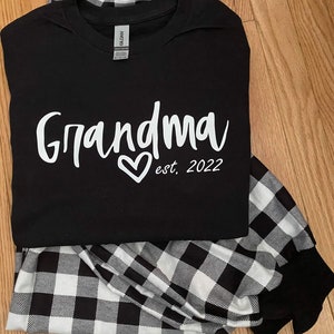 K&ouml;nnte beinhalten: Schwarz-wei&szlig;e Buffalo-Karos Schlafanzughose und ein schwarzes T-Shirt mit dem Text "Grandma est. 2022" in Wei&szlig;.