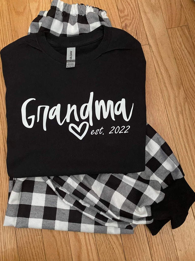 K&ouml;nnte beinhalten: Schwarz-wei&szlig;e Buffalo-Karos Schlafanzughose und ein schwarzes T-Shirt mit dem Text "Grandma est. 2022" in Wei&szlig;.