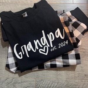 K&ouml;nnte beinhalten: Schwarz-wei&szlig;e karierte Pyjamahose mit einem schwarzen T-Shirt, auf dem in wei&szlig;er Schrift "Grandpa est. 2024" mit einem Herz steht.