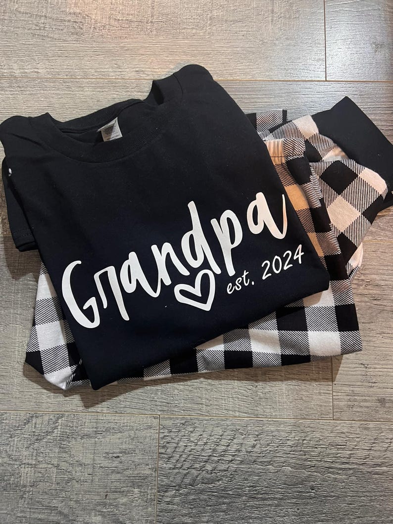 K&ouml;nnte beinhalten: Schwarz-wei&szlig;e karierte Pyjamahose mit einem schwarzen T-Shirt, auf dem in wei&szlig;er Schrift "Grandpa est. 2024" mit einem Herz steht.