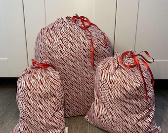 May include: Three red and white striped fabric gift bags with red ribbon ties. The bags are arranged in a pyramid shape, with the largest bag at the bottom and the smallest bag at the top.