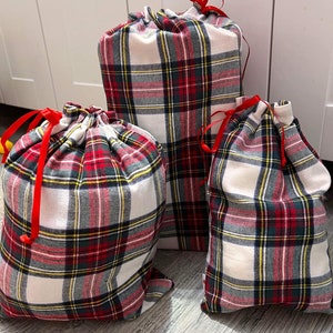 May include: Three red and white tartan gift bags with red ribbon ties. The bags are made of fabric and have a drawstring closure.