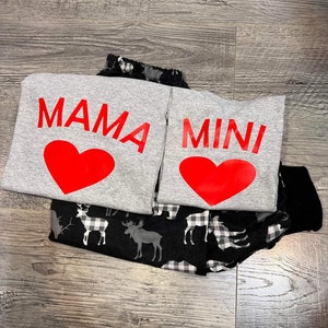 May include: Two gray sweatpants with red hearts and the words "MAMA" and "MINI" printed on them. The sweatpants are folded on top of a black and white plaid fabric with a moose and deer design.