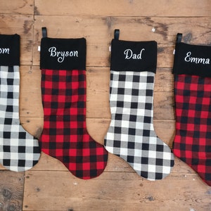 May include: Four red and black buffalo check Christmas stockings with black felt cuffs. The cuffs are embroidered with the names "Mom", "Bryson", "Dad", and "Emma".