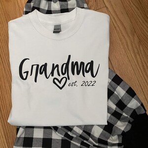 K&ouml;nnte beinhalten: Wei&szlig;es T-Shirt mit schwarzem Text, der "Grandma est. 2022" mit einem Herzen sagt. Das Shirt wird mit einem schwarz-wei&szlig; karierten Schlafanzug kombiniert.