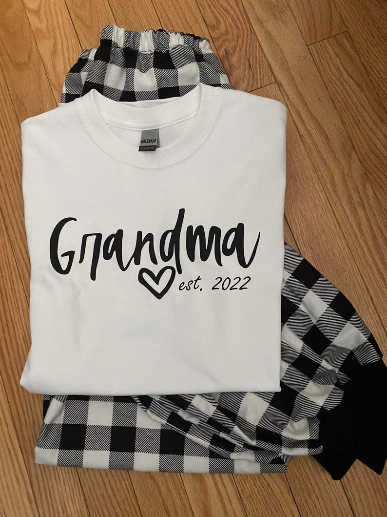 K&ouml;nnte beinhalten: Wei&szlig;es T-Shirt mit schwarzem Text, der "Grandma est. 2022" mit einem Herzen sagt. Das Shirt wird mit einem schwarz-wei&szlig; karierten Schlafanzug kombiniert.