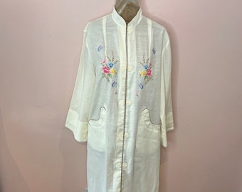 May include: Cream-colored robe with floral embroidery. The robe features a mandarin collar, long sleeves, and a button-up front. The embroidery includes pink, blue, and yellow flowers. The robe is displayed on a mannequin with a stand.