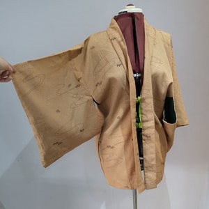 Puede incluir: Un kimono japonés de color beige con un patrón de ondas y detalles marrones. El kimono se muestra en un maniquí.