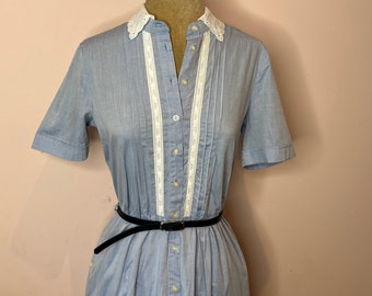 May include: A light blue button-down dress with a white lace collar and trim. The dress has a fitted bodice and a flowing skirt. It is belted with a black belt.
