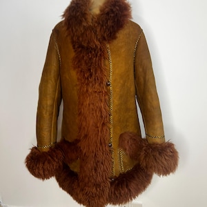 May include: A vintage brown shearling coat with a shaggy brown fur collar, cuffs, and front panel. The coat has a button-up front and gold stitching details. The coat is displayed on a mannequin.