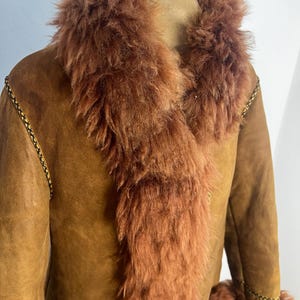 May include: A brown suede coat with a fluffy, reddish-brown fur collar and trim. The coat has a classic design with a tailored fit, and the fur adds a touch of luxury. The coat is displayed on a mannequin.