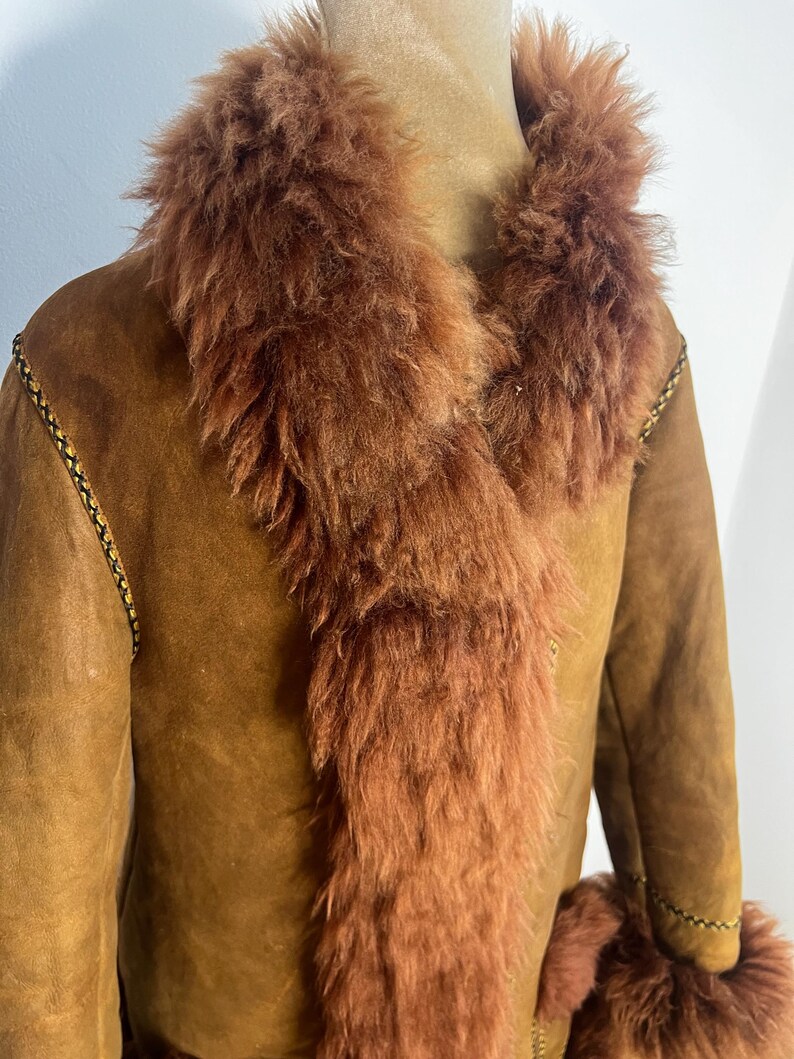 May include: A brown suede coat with a fluffy, reddish-brown fur collar and trim. The coat has a classic design with a tailored fit, and the fur adds a touch of luxury. The coat is displayed on a mannequin.