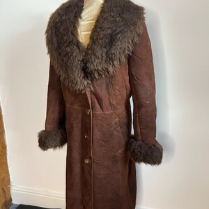 May include: A long, brown suede coat with a fur collar and cuffs. The coat has a button-down front and is displayed on a mannequin. The fur is a darker shade of brown than the coat.
