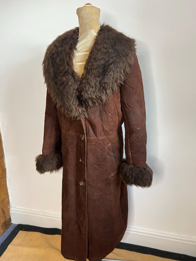 May include: A long, brown suede coat with a fur collar and cuffs. The coat has a button-down front and is displayed on a mannequin. The fur is a darker shade of brown than the coat.