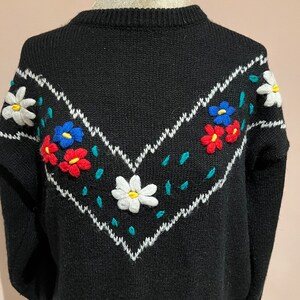 May include: Black knitted sweater with a white, blue, red, and green embroidered floral design on the front.
