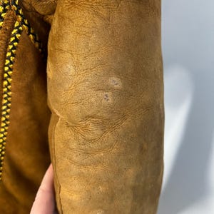 May include: Close-up of a tan leather jacket sleeve. The sleeve has a textured surface with visible wear and small imperfections. A decorative seam with black and yellow stitching runs along the edge of the jacket.