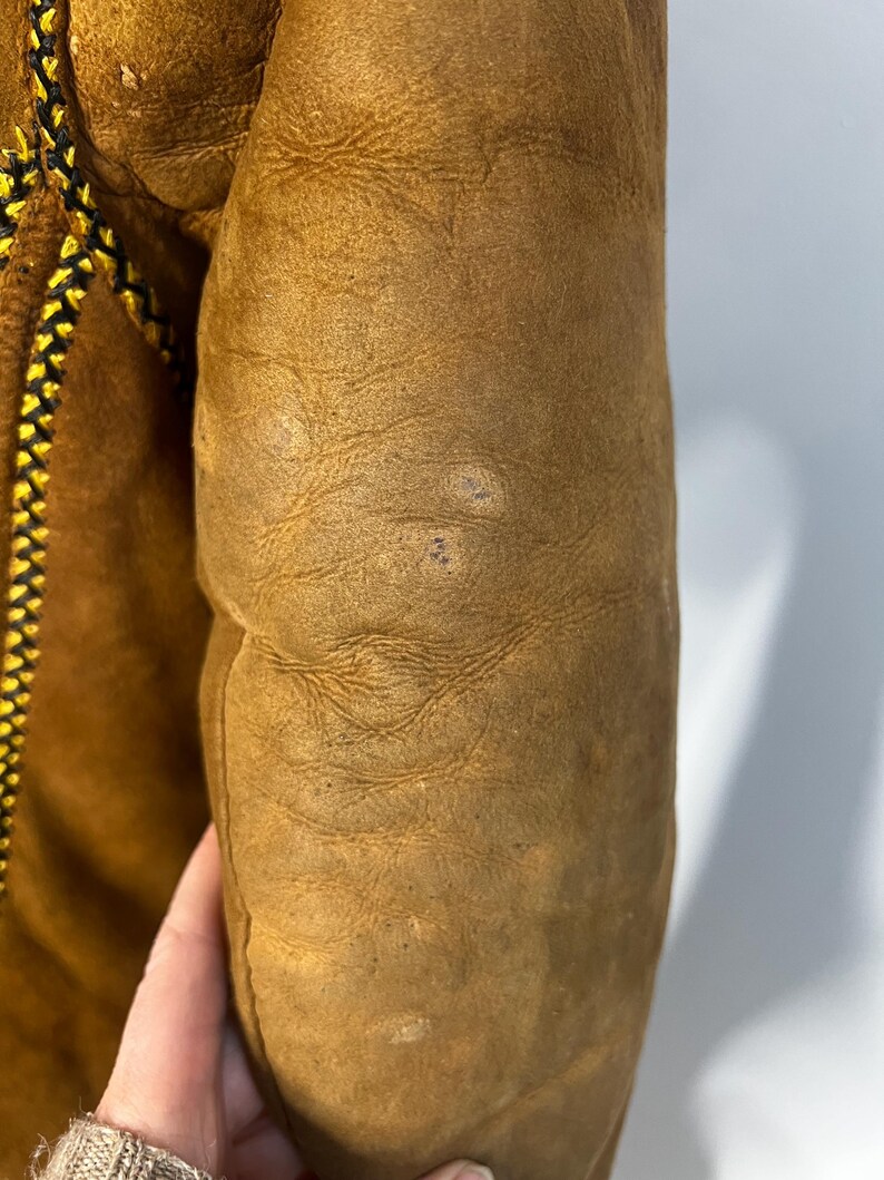 May include: Close-up of a tan leather jacket sleeve. The sleeve has a textured surface with visible wear and small imperfections. A decorative seam with black and yellow stitching runs along the edge of the jacket.