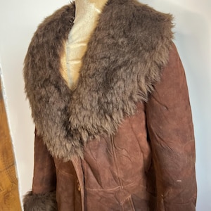 May include: A brown suede coat with a faux fur collar and cuffs. The coat is buttoned up and appears to be vintage. The fur is a darker shade of brown than the suede. The coat is displayed on a mannequin.