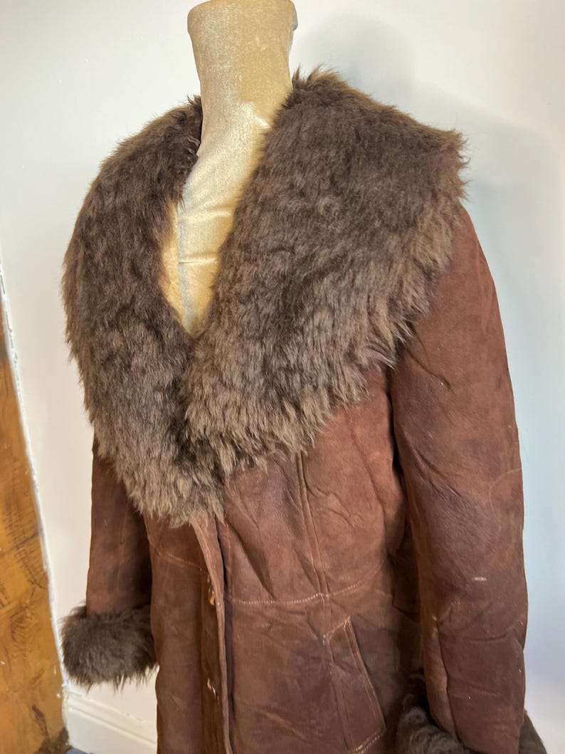 May include: A brown suede coat with a faux fur collar and cuffs. The coat is buttoned up and appears to be vintage. The fur is a darker shade of brown than the suede. The coat is displayed on a mannequin.
